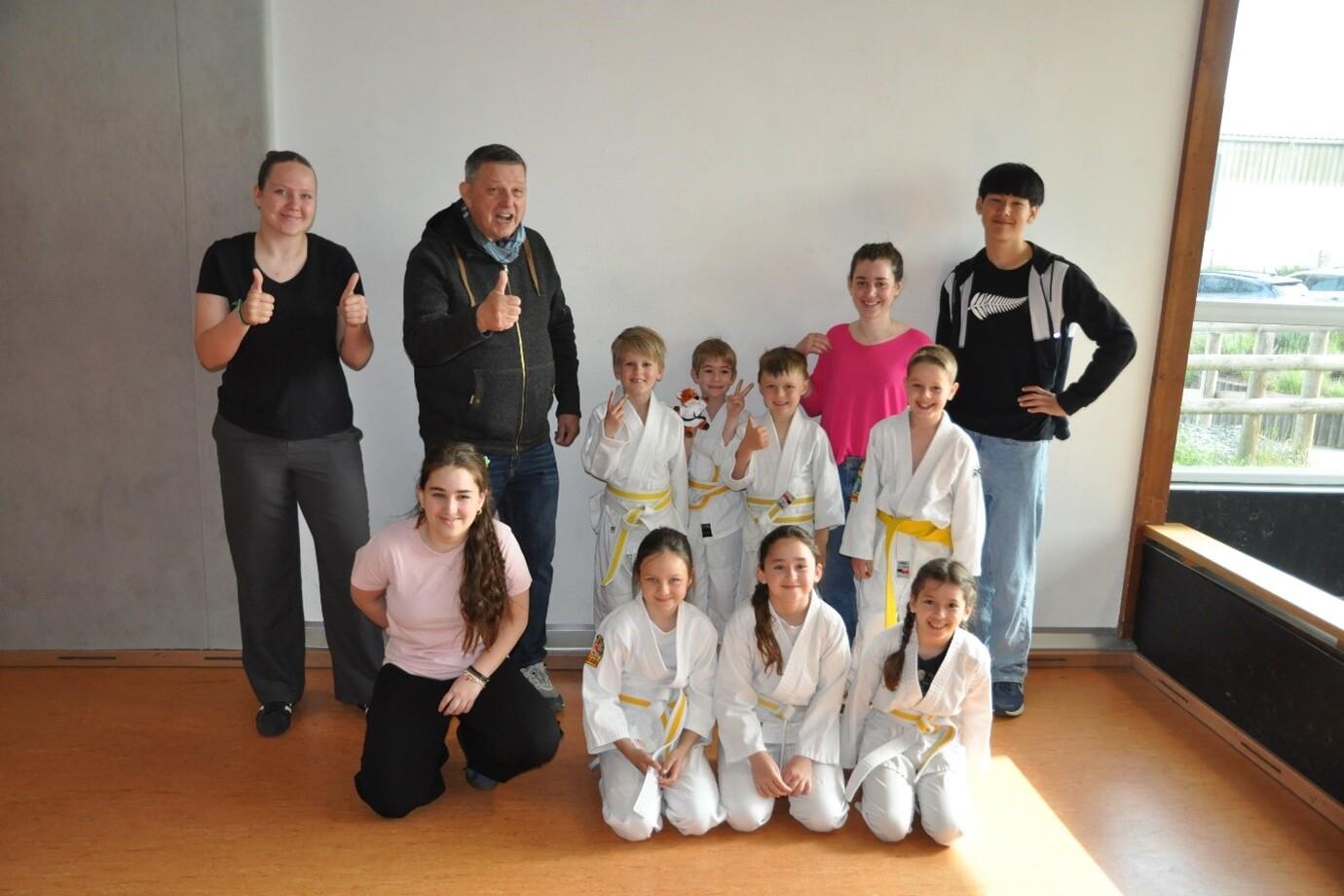 Gruppe von zehn Kindern und Erwachsenen in einem Raum, alle lächeln und zeigen Daumen hoch; einige tragen Karateanzüge.