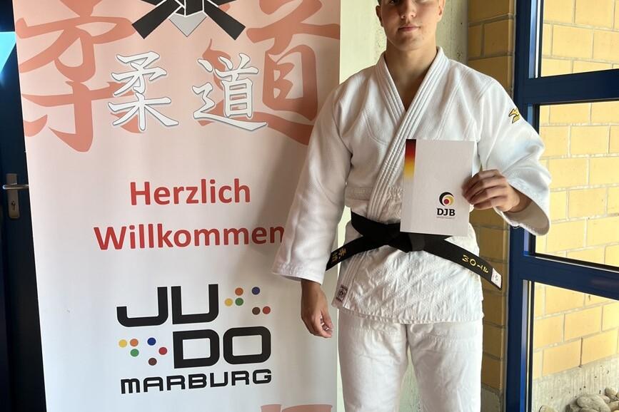 Junger Judoka in weißem Gi und schwarzem Gürtel hält ein Zertifikat vor einem Judo-Verband-Banner in Marburg.