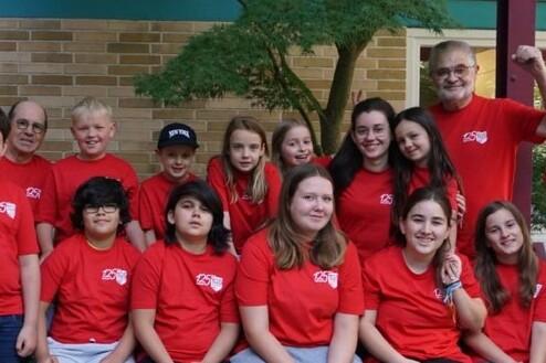 Gruppe von 15 Personen in roten T-Shirts, stehend und sitzend, vor einer Mauer mit Pflanzen im Hintergrund.
