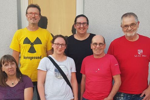 Gruppenfoto von sechs Personen vor einer grauen Wand, alle lächeln und tragen unterschiedliche Kleidung.