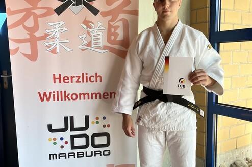 Junger Judoka in weißem Gi und schwarzem Gürtel hält ein Zertifikat vor einem Judo-Verband-Banner in Marburg.