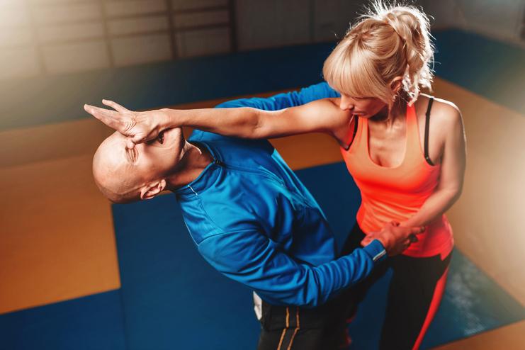 Eine Frau in Sportbekleidung übt einen Selbstverteidigungsgriff an einem Mann in blauer Kleidung auf einer Mattenfläche aus.