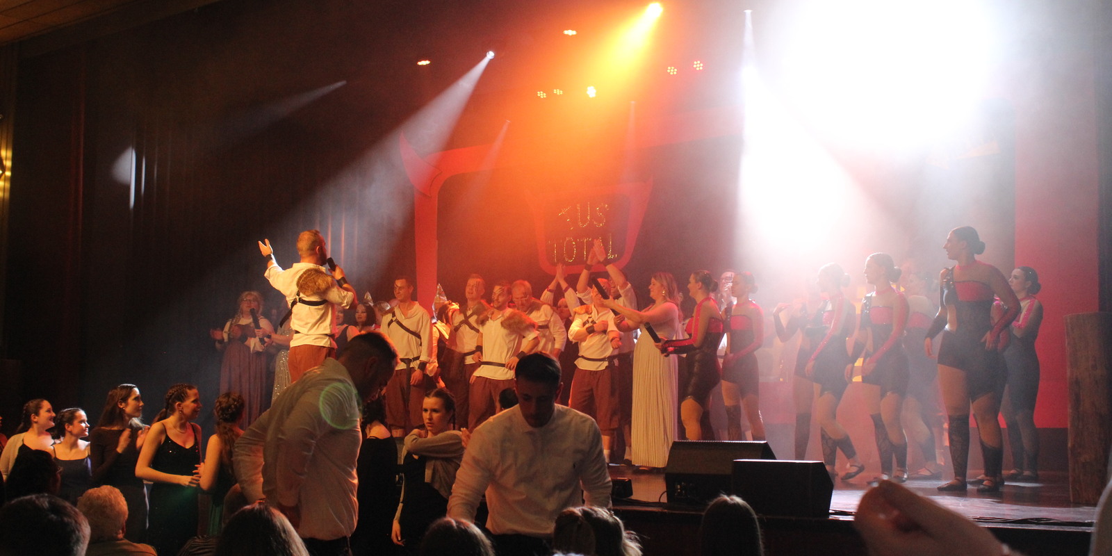 B&uuml;hne mit mehreren T&auml;nzern und S&auml;ngerinnen in Kost&uuml;men, beleuchtet von Scheinwerfern, w&auml;hrend das Publikum zuschaut.