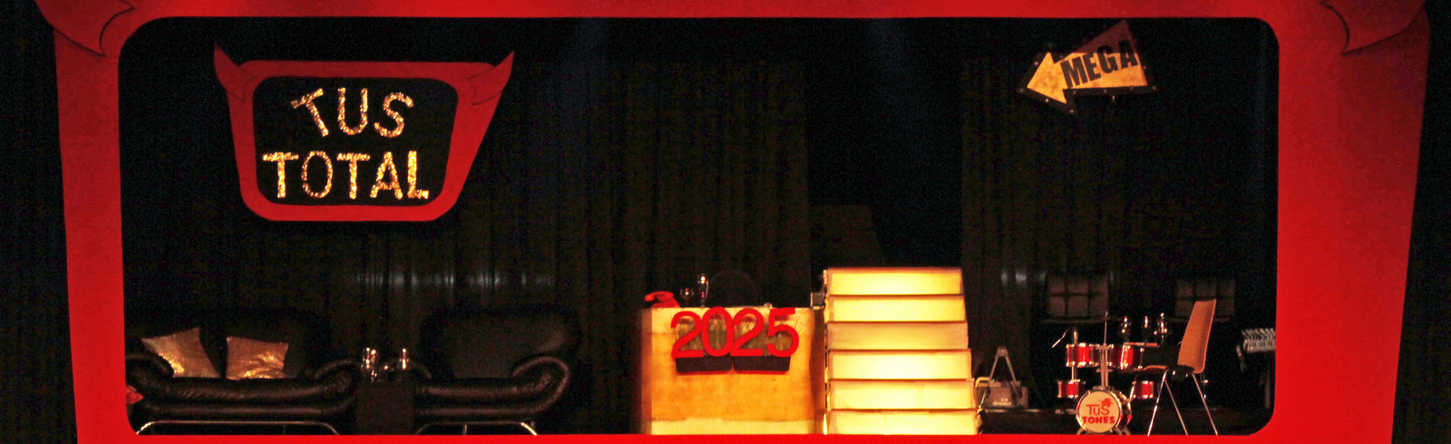 B&uuml;hnenbild mit rotem Rahmen, Schriftzug "KUS TOTAL", Couch, B&uuml;hne mit Stufen und Schlagzeug im Hintergrund.