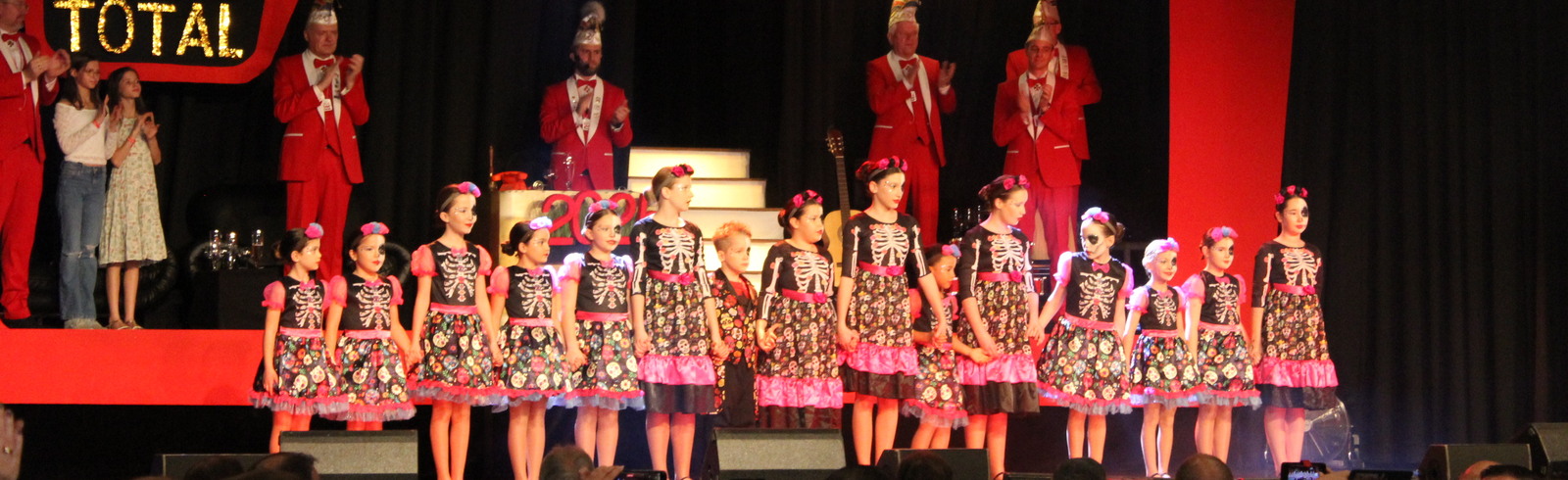 Kinder in bunten Kleidern mit Skelett-Muster stehen auf der B&uuml;hne vor applaudierendem Publikum und festlicher Dekoration.