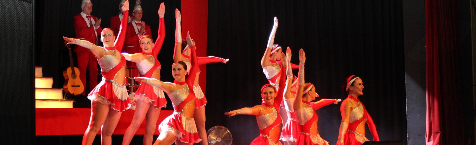 Tanzgruppe in roten Kost&uuml;men f&uuml;hrt eine Choreografie auf einer B&uuml;hne mit B&uuml;hnenbild und Musikern im Hintergrund auf.