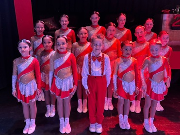 Tanzgruppe in roten Kostümen steht vor einem Banner mit der Aufschrift "TUS TOTAL" auf einer Bühne.