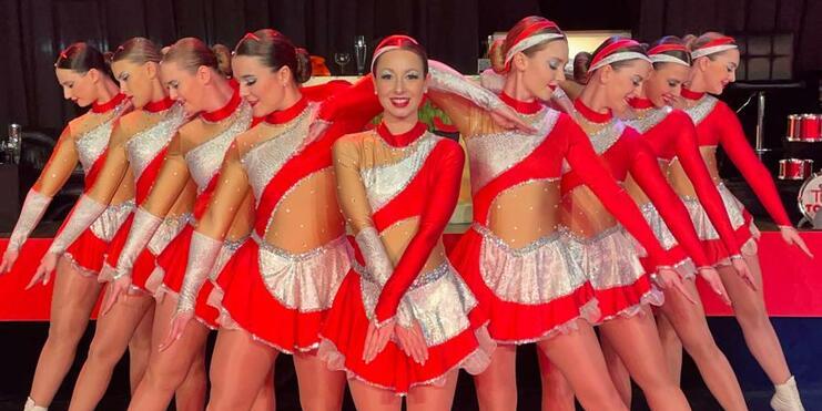 Gruppe von Tänzerinnen in roten und silbernen Kostümen posiert synchron auf einer Bühne mit farbigem Licht.