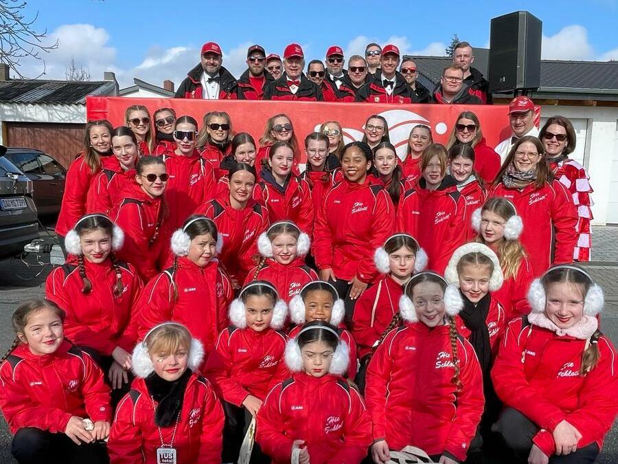 Gruppe von Mädchen und Erwachsenen in roten Jacken mit Mützen, vor einem Banner und blauem Himmel.