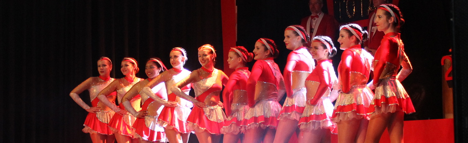 Gruppe von T&auml;nzerinnen in rot-wei&szlig;en Kost&uuml;men auf einer B&uuml;hne mit Scheinwerfern und B&uuml;hnenkulisse im Hintergrund.