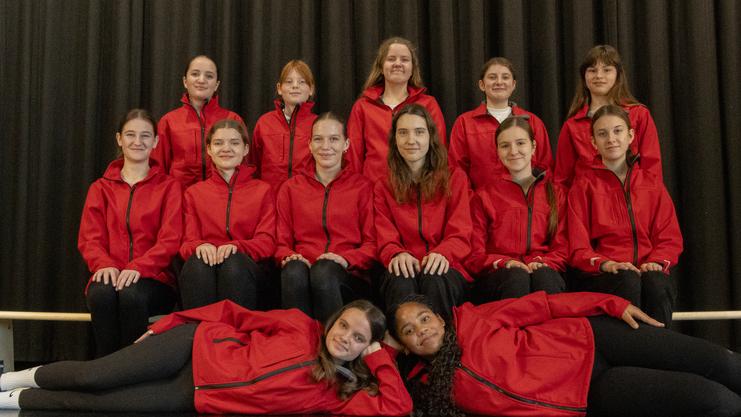 Gruppe von 14 Mädchen in roten Jacken, posierend auf einer Bank vor einem schwarzen Vorhang.