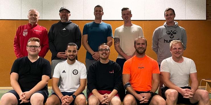 Gruppe von zehn Männern, sitzend auf einer Bank in Turnkleidung, in einem Sportraum mit grünem Boden.