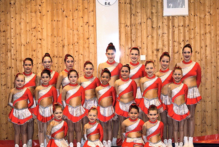 Gruppe von 16 jungen T&auml;nzerinnen in orange-roten Kost&uuml;men, aufgestellt vor einer Holzwand in einer Sporthalle.