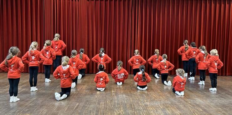 Gruppe von Mädchen in roten Trainingsjacken, die auf einer Holzfläche mit rotem Vorhang hinter ihnen posieren.