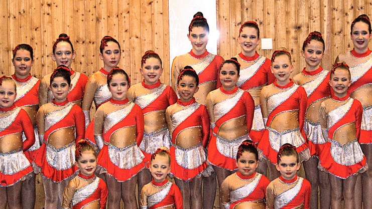 Gruppe von 16 jungen T&auml;nzerinnen in orange-roten Kost&uuml;men, aufgestellt vor einer Holzwand in einer Sporthalle.