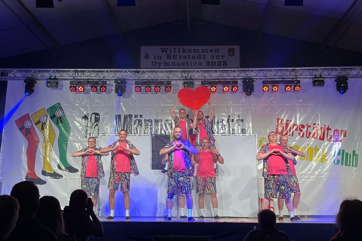 Tanzgruppe auf B&uuml;hne in bunten Kost&uuml;men, mit Herz-Requisiten, vor einem Banner mit dem Text "Willkommen zur Gymnastica 2023".