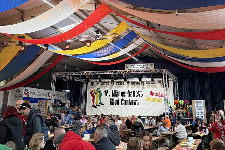 Blick in eine Festhalle mit bunten, gewellten Stoffbahnen und Tischen, an denen Besucher an einem Wettbewerb teilnehmen.