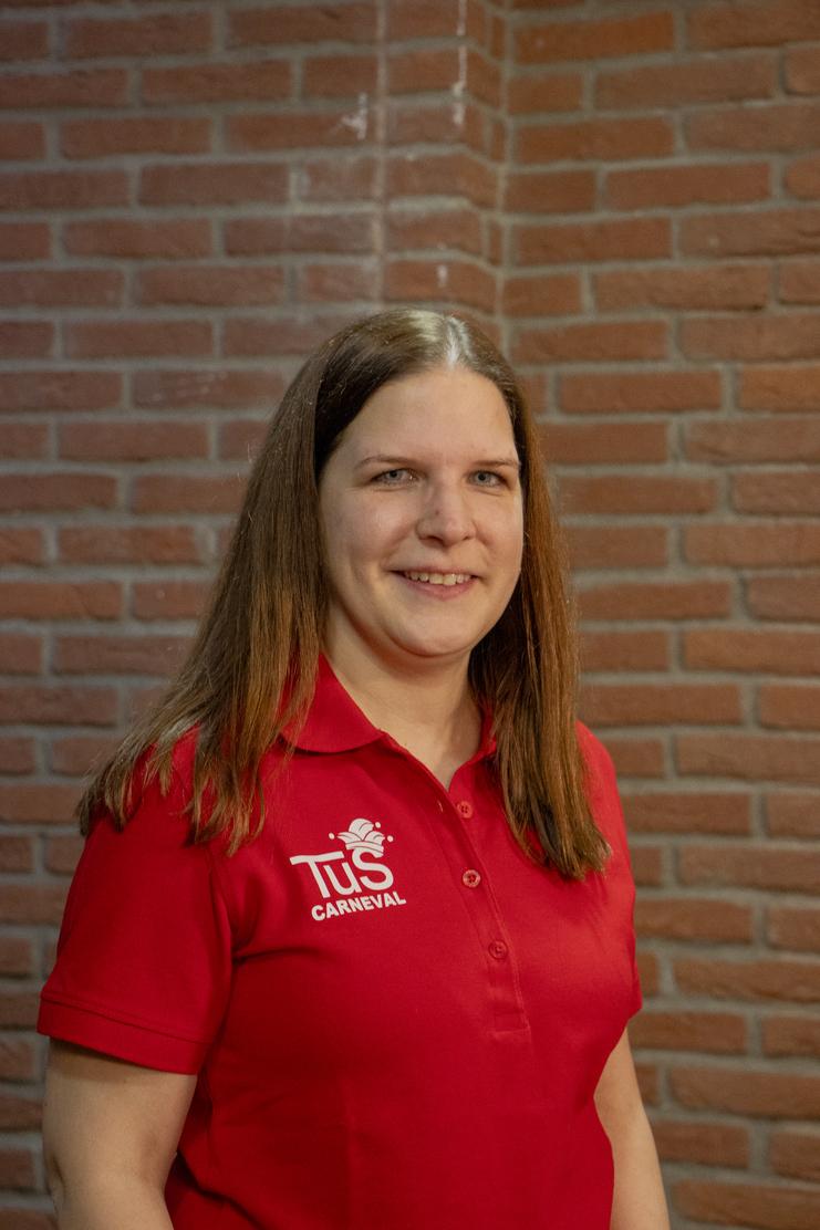 Frau mit langen, braunen Haaren trägt ein rotes Poloshirt mit „TuS Carneval“-Logo vor einer Ziegelwand.