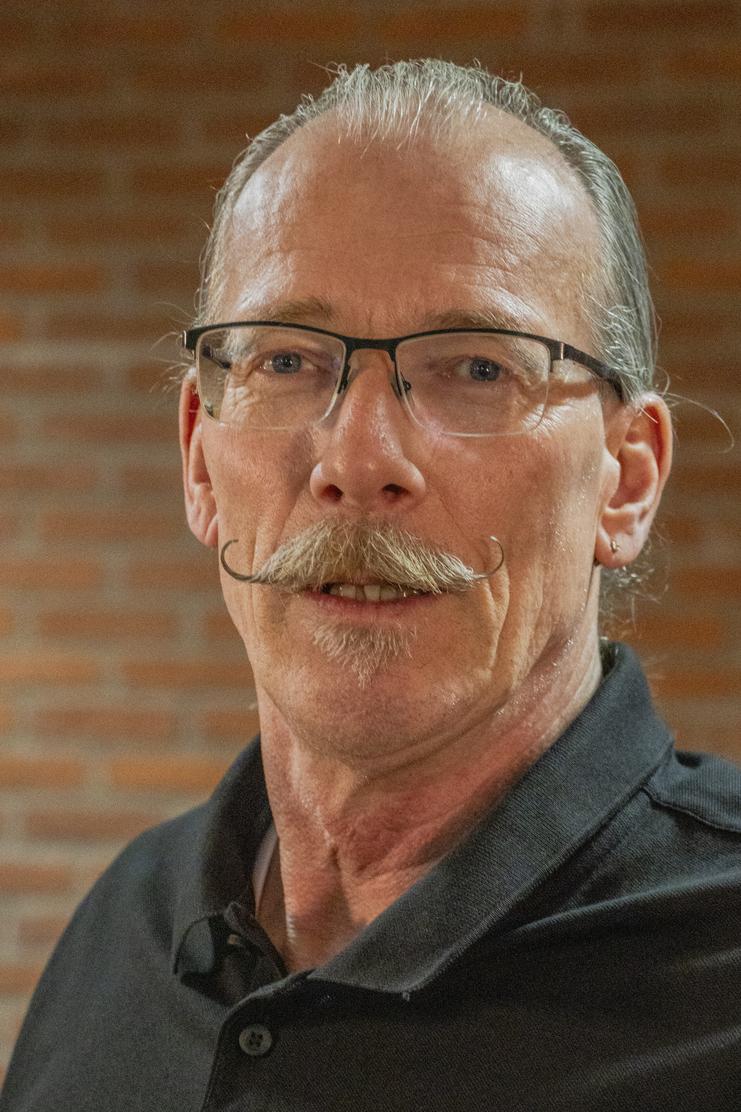 Mann mit Brille und großem Schnurrbart trägt ein schwarzes Hemd vor einem Ziegelwand-Hintergrund.