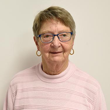 Porträt einer älteren Frau mit Brille, in einem rosa Pullover, vor hellem Hintergrund. Sie lächelt leicht.