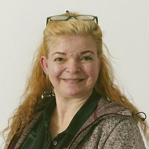 Frau mit blonden, lockigen Haaren und Brille lächelt vor einem neutralen Hintergrund.