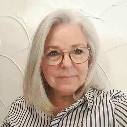 Porträt einer Frau mit grauen Haaren und Brille, vor einer strukturierten weißen Wand mit Wellenmuster.