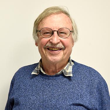 Portrait eines lächelnden älteren Mannes mit Brille, tragt einen blauen Pullover und ein kariertes Hemd. Hintergrund hell.