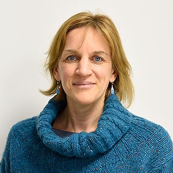 Porträt einer lächelnden Frau mit blonden Haaren, die einen blauen Pullover trägt, vor einem grauen Hintergrund.