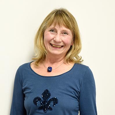 Lächelnde Frau mit blonden Haaren trägt ein blaues Oberteil mit Glitzerverzierung vor einer hellen Wand.