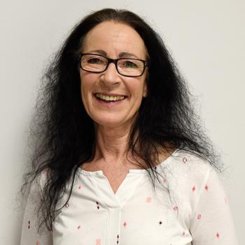 Lächelnde Frau mit langen, schwarzen Haaren und Brille, steht vor einem hellen Hintergrund, trägt ein weißes Oberteil.