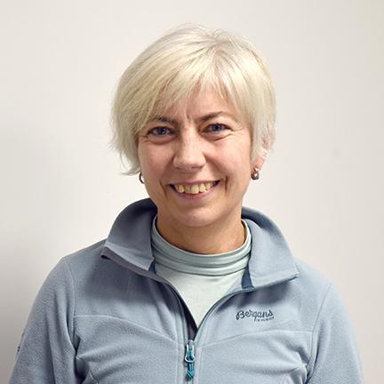 Porträt einer lächelnden Frau mit blonden, kurzen Haaren, die einen hellblauen Fleece-Pullover trägt. Hintergrund neutral.