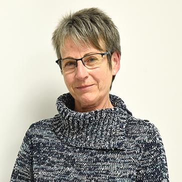 Frau mit kurzen grauen Haaren und Brille, trägt einen graublauen Pullover, steht vor einer neutralen Wand.