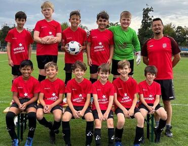 Gruppe junger Fußballspieler in roten Trikots, stehend und sitzend auf einer Wiese mit einem Ball.