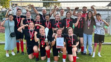 Gruppenfoto einer Fußballmannschaft mit Medaillen, Pokal und Urkunde, umgeben von Trainer und Unterstützern auf dem Spielfeld.