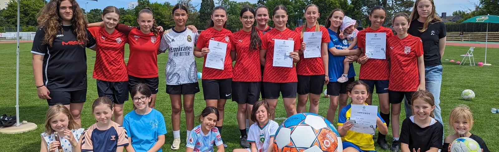 Gruppe von M&auml;dchen in Fu&szlig;balltrikots mit Urkunden und einem gro&szlig;en Fu&szlig;ball auf einem gr&uuml;nen Sportplatz.
