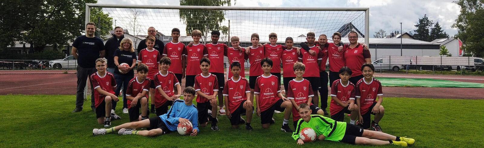 Gruppe von Jugendlichen und Trainern in roten Trikots auf einem Fu&szlig;ballplatz, vor einem Tor stehend.