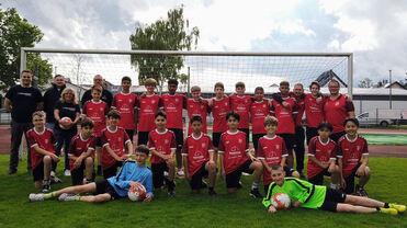 Gruppe von Jugendlichen und Trainern in roten Trikots auf einem Fußballplatz, vor einem Tor stehend.