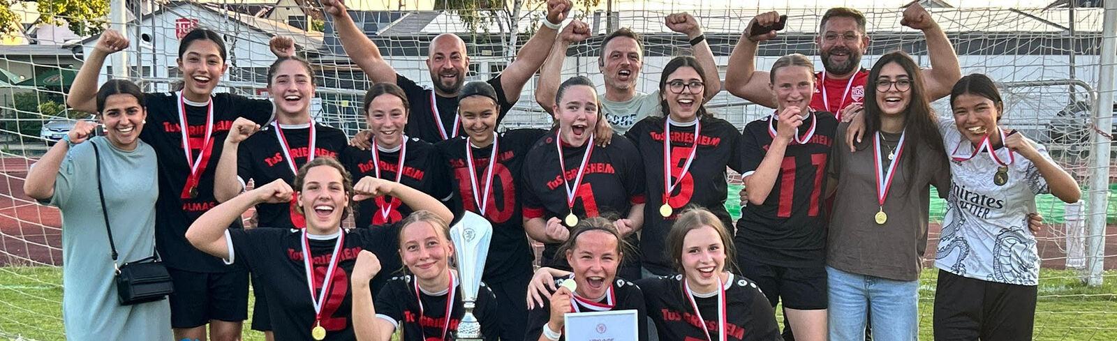 Gruppenfoto einer Fu&szlig;ballmannschaft mit Medaillen, Pokal und Urkunde, umgeben von Trainer und Unterst&uuml;tzern auf dem Spielfeld.