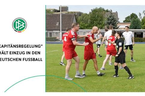 Fußballtrainer diskutieren mit Schiedsrichterin auf dem Spielfeld, während Spieler im Hintergrund stehen.