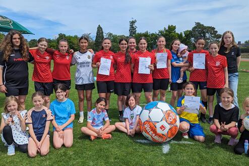 Gruppe von Mädchen in Fußballtrikots mit Urkunden und einem großen Fußball auf einem grünen Sportplatz.