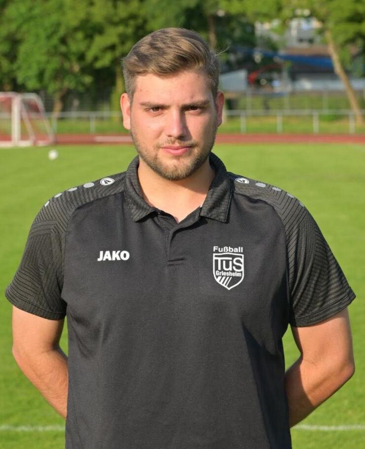Ein junger Mann steht mit verschränkten Armen auf einem Fußballplatz, im Hintergrund sind Tore und Bäume sichtbar.