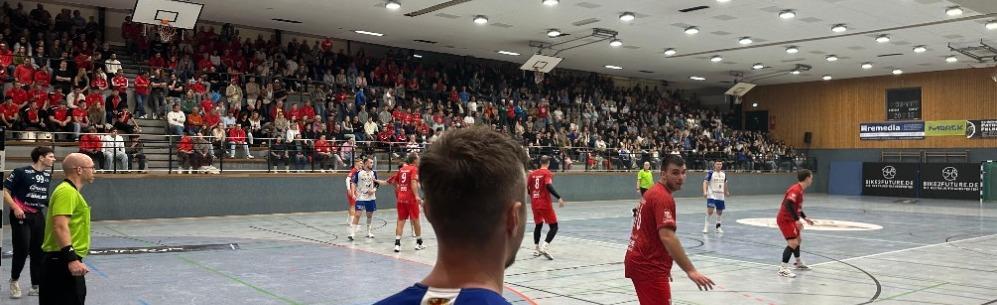 Handballspiel in einer vollbesetzten Halle mit Spielern in roten und blauen Trikots. Zuschauer feuern enthusiastisch an.