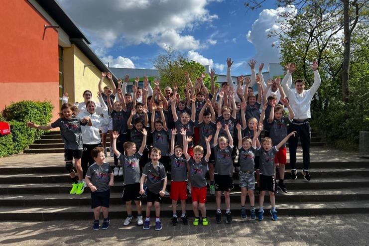 Gruppenshot von Kindern und Betreuern, die auf Treppen stehen und jubelnd die Hände heben, unter blauem Himmel.