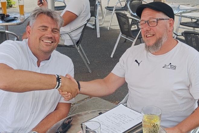 Zwei Männer schütteln sich die Hände am Tisch in einem Außenbereich eines Cafés, im Hintergrund sind andere Gäste sichtbar.