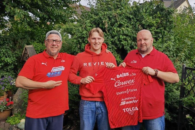 Drei Männer in roten T-Shirts posieren im Freien mit einem Trikot vor einem Garten.