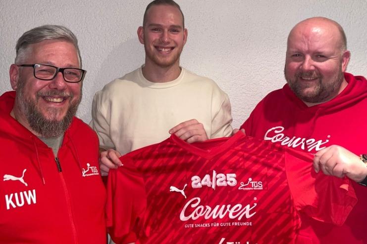 Drei Männer halten ein rotes Fußballtrikot mit Sponsorenlogos vor einer hellen Wand.
