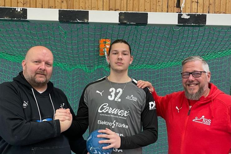 Drei Personen stehen vor einem Handballtor, ein Spieler hält einen Handball, während die anderen ihn umarmen.