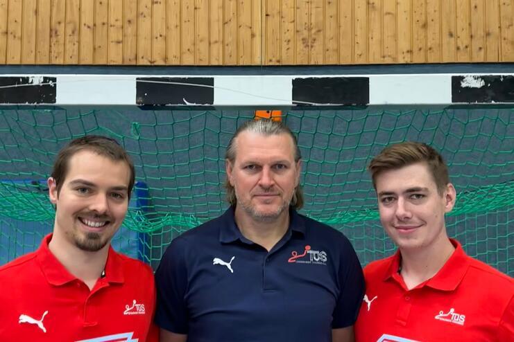 Drei Personen stehen vor einem grünen Handballtor, tragen sportliche Kleidung und lächeln in die Kamera.