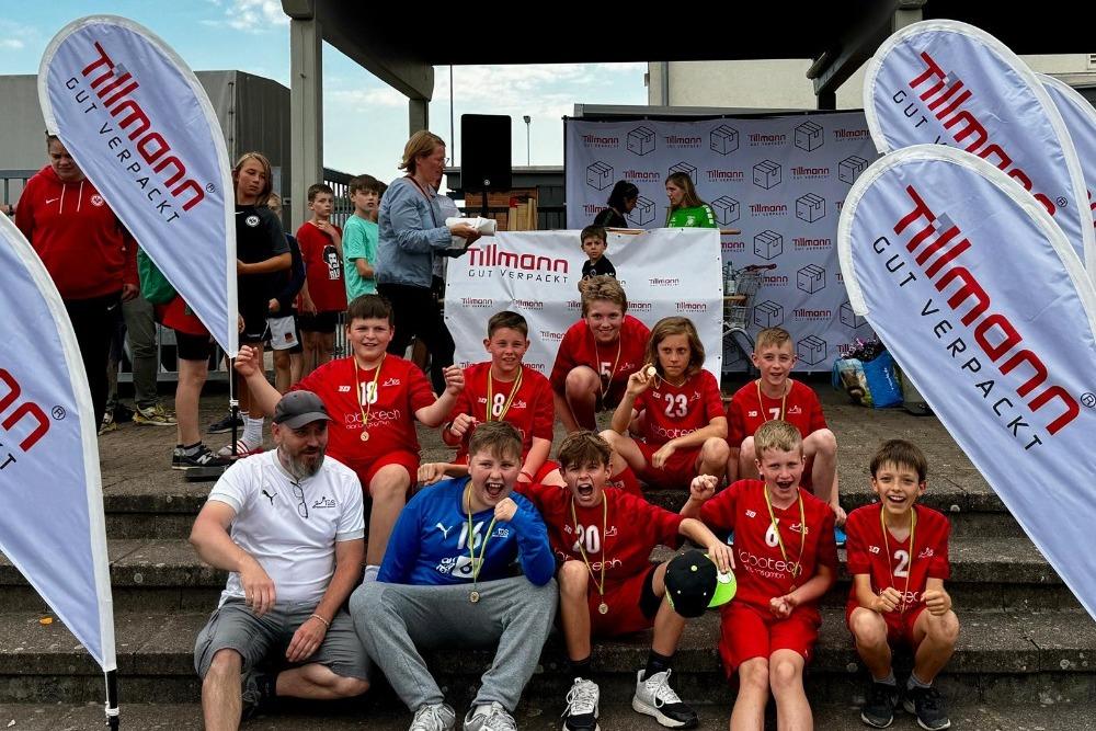 Gruppe von Kindern in roten Sporttrikots mit Medaillen sitzt auf Stufen, umgeben von Fahnen und Zuschauern.