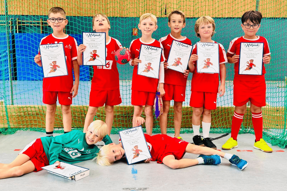Gruppenfoto von acht Kindern in roten Sportanzügen, die stolz Urkunden in einer Sporthalle zeigen. Ein Kind liegt lächelnd auf dem Boden.